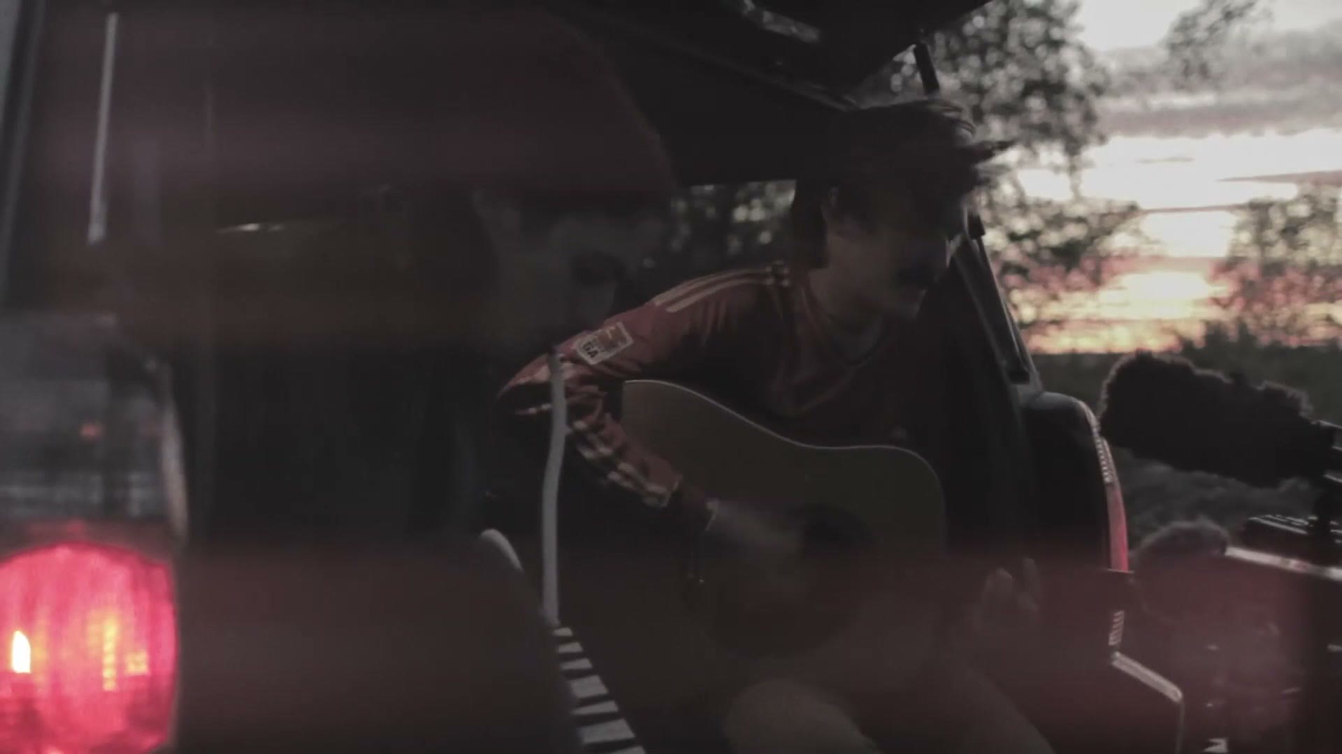 A wide shot of a musician playing acoustic guitar in the open bed of a pickup truck, silhouetted against an orange sunset