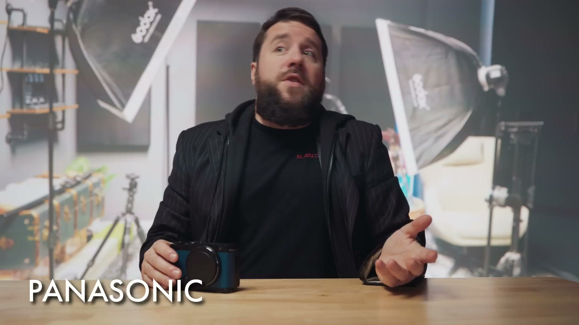 Matt dressed as Panasonic in a black jacket with a small camera on the desk in front of him