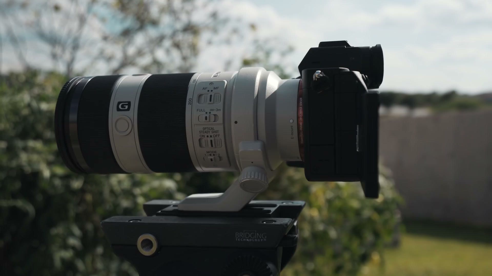A Sony a7S III on a tripod outdoors with a white Sony 70-200mm G lens mounted by its own lens ring, leaving air free to move around the camera body