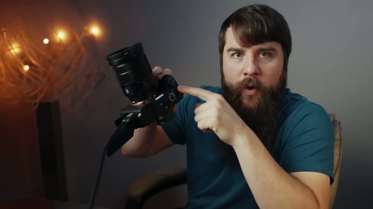 Matt talking to camera pointing at the shutter button on the a7S III, string lights glowing behind him