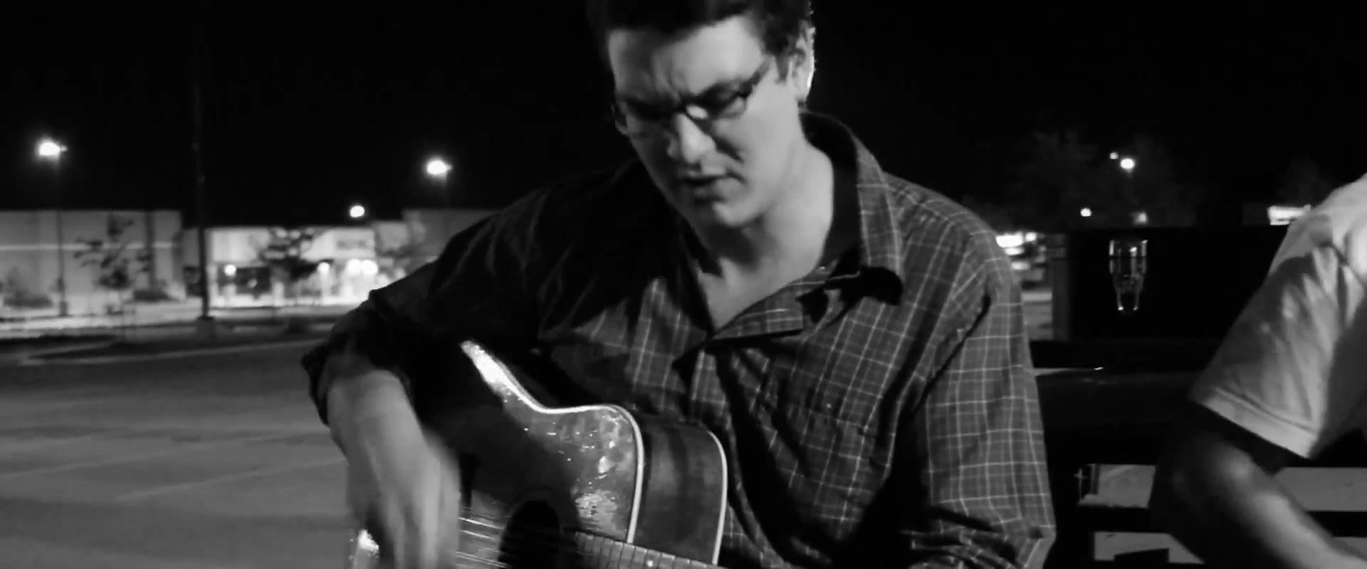 Black and white close-up of Curt Vernon looking down at his guitar strings, storefronts lit up across the empty lot