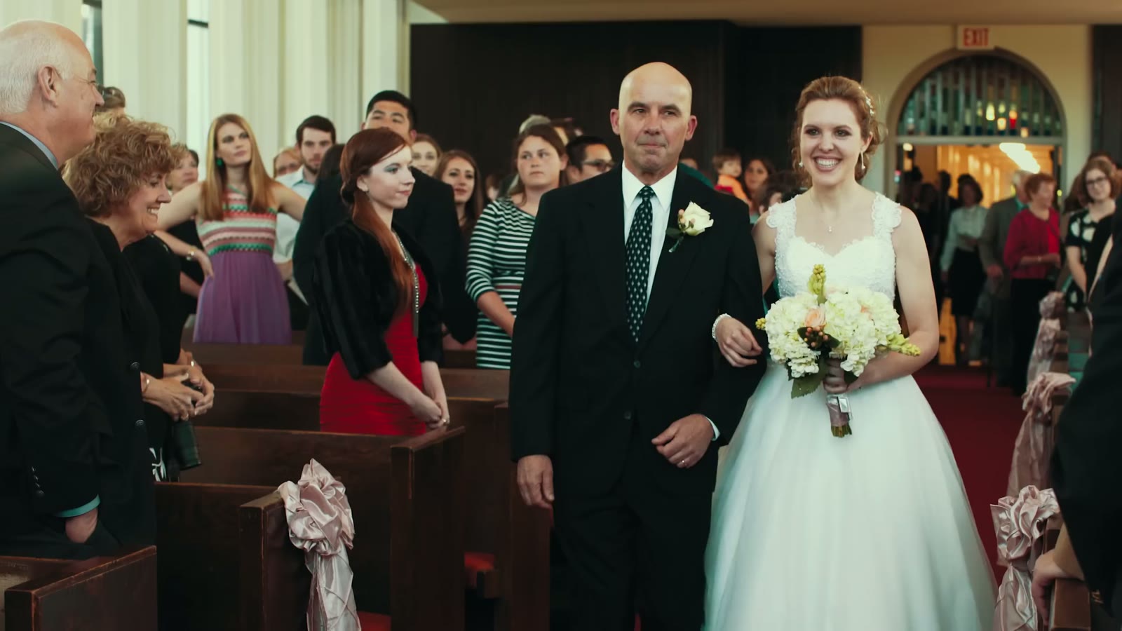Bride in a strapless gown walking down the aisle on her father's arm, guests standing in the pews