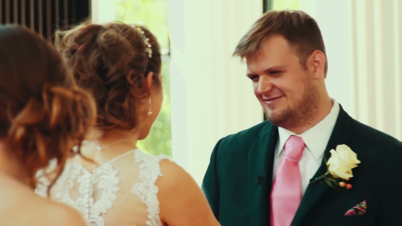 Groom in a green jacket and pink tie smiling at a different bride in a lace-back gown during vows