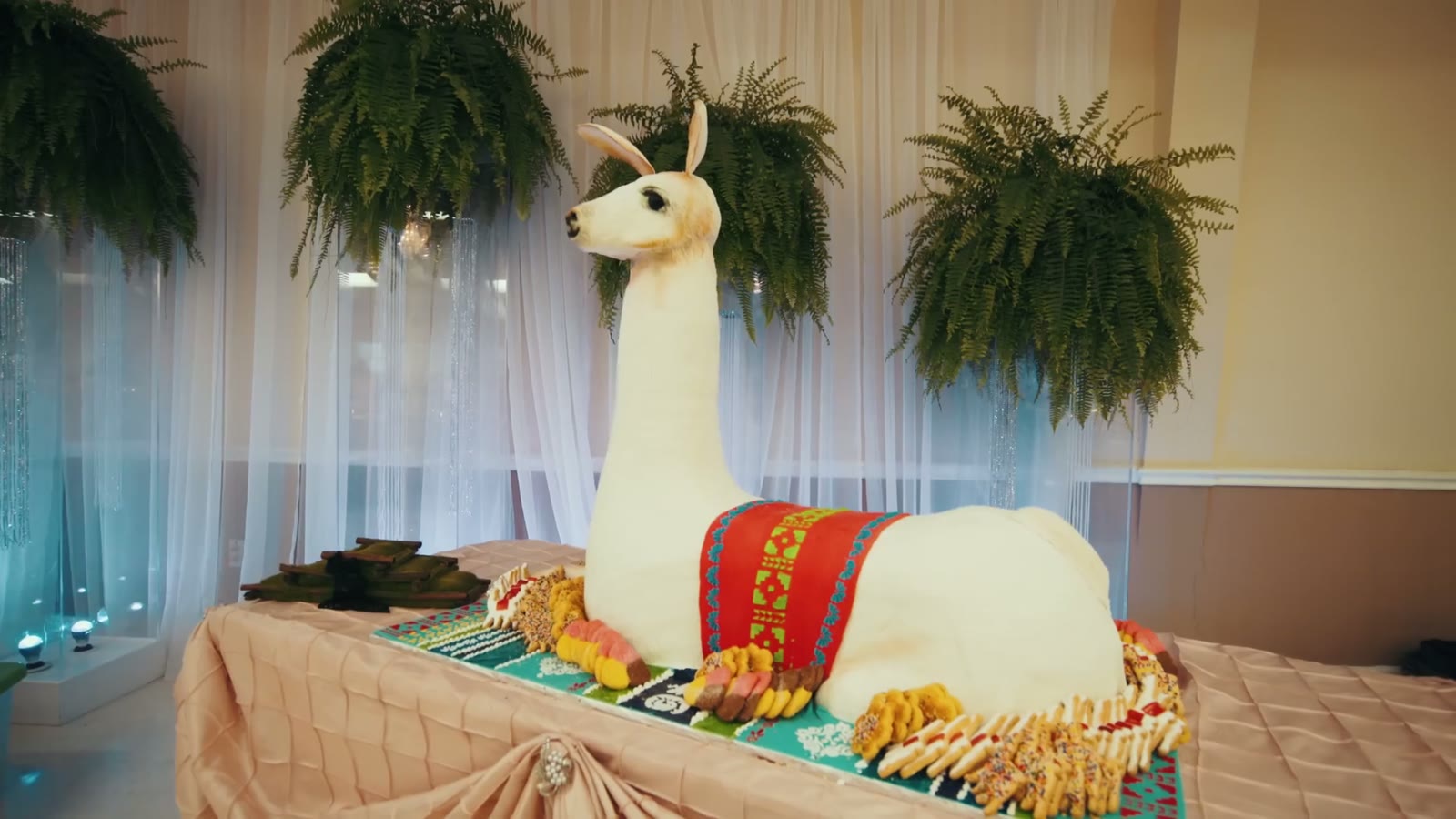 Life-size llama-shaped wedding cake draped in a colorful blanket, set on a dessert table with ferns