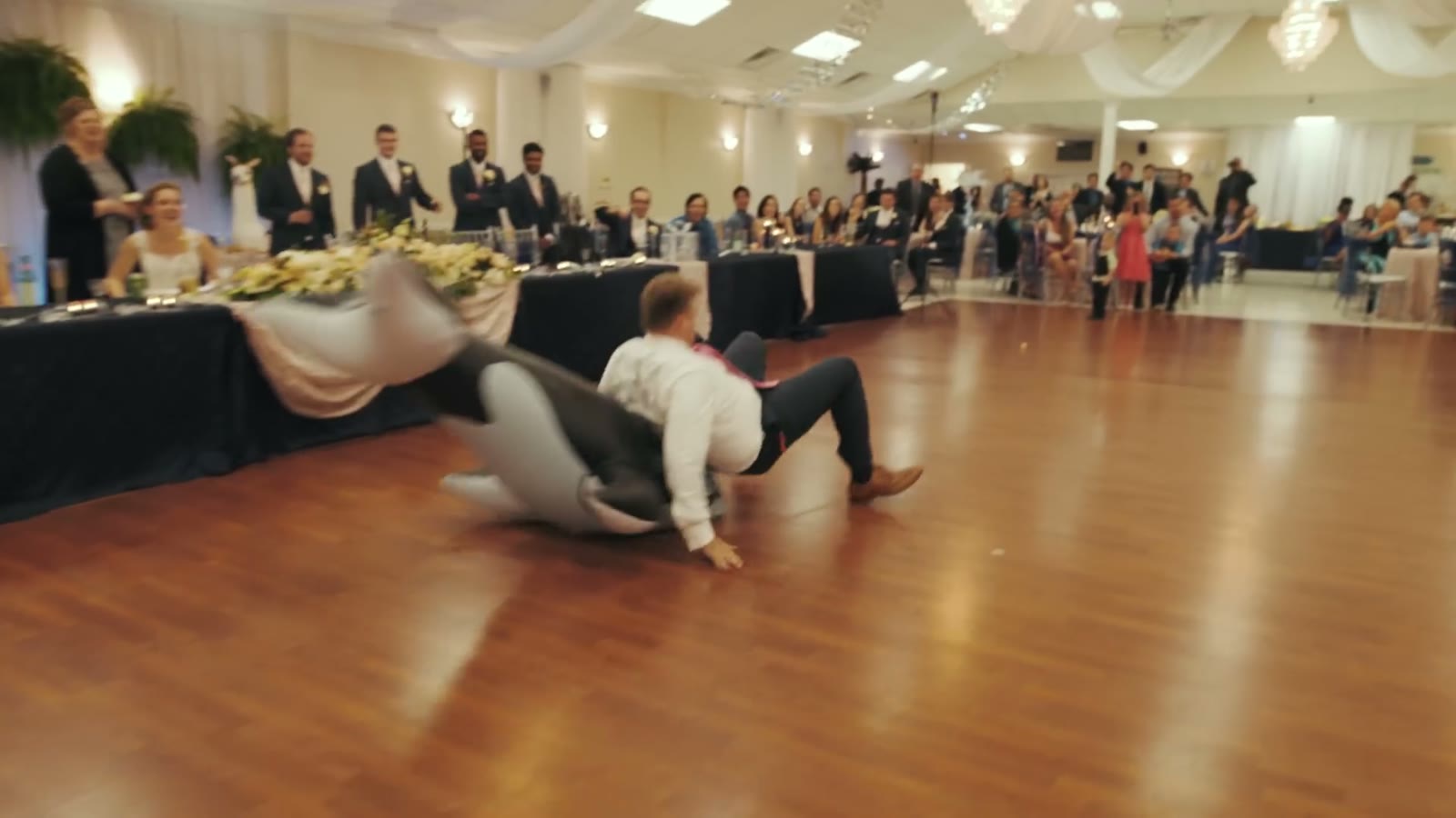 Groom wrestling and dancing on the floor with a person in an inflatable whale costume as guests watch