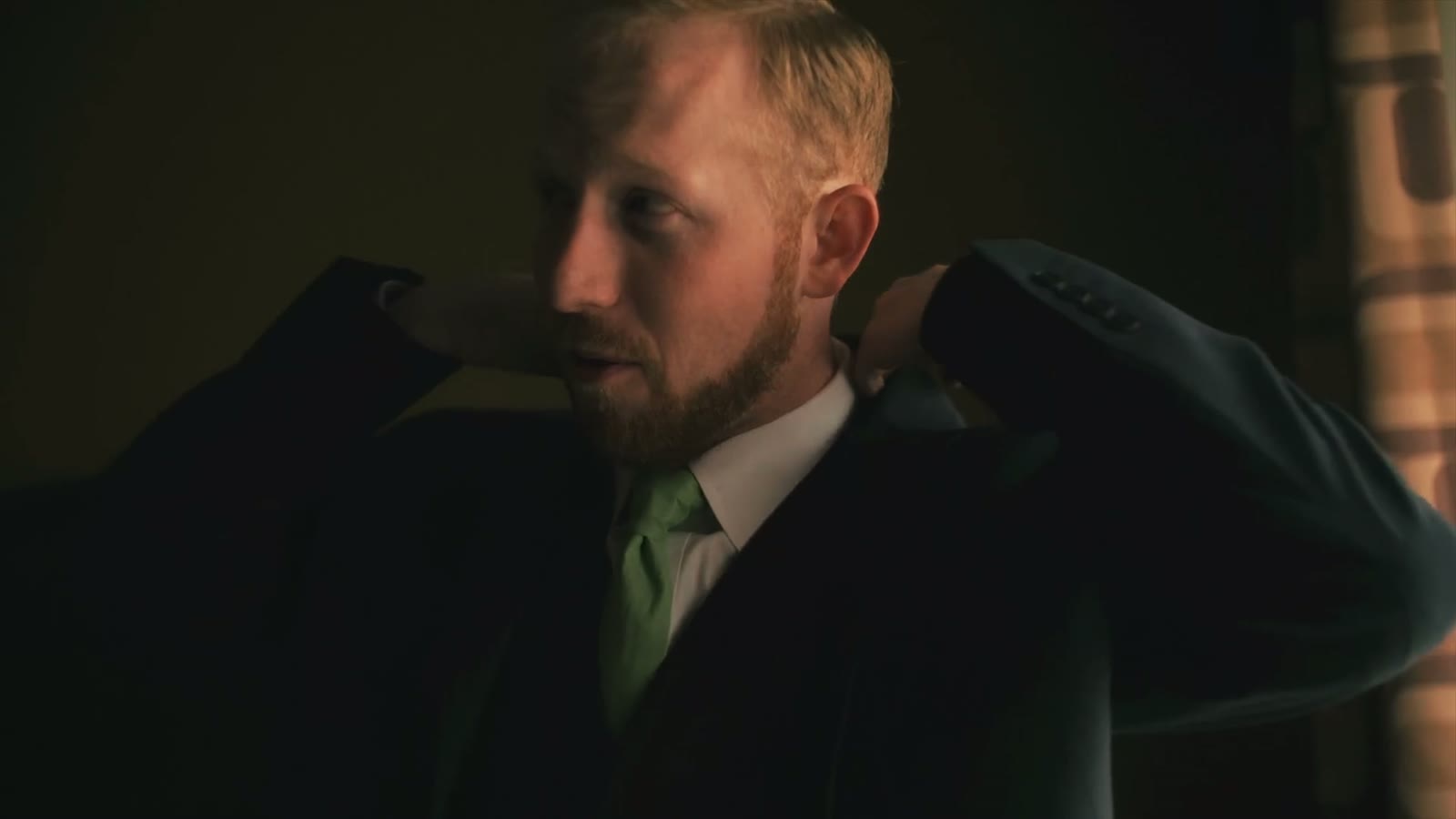 Groom adjusting his jacket lapels in low warm light, wearing a green tie