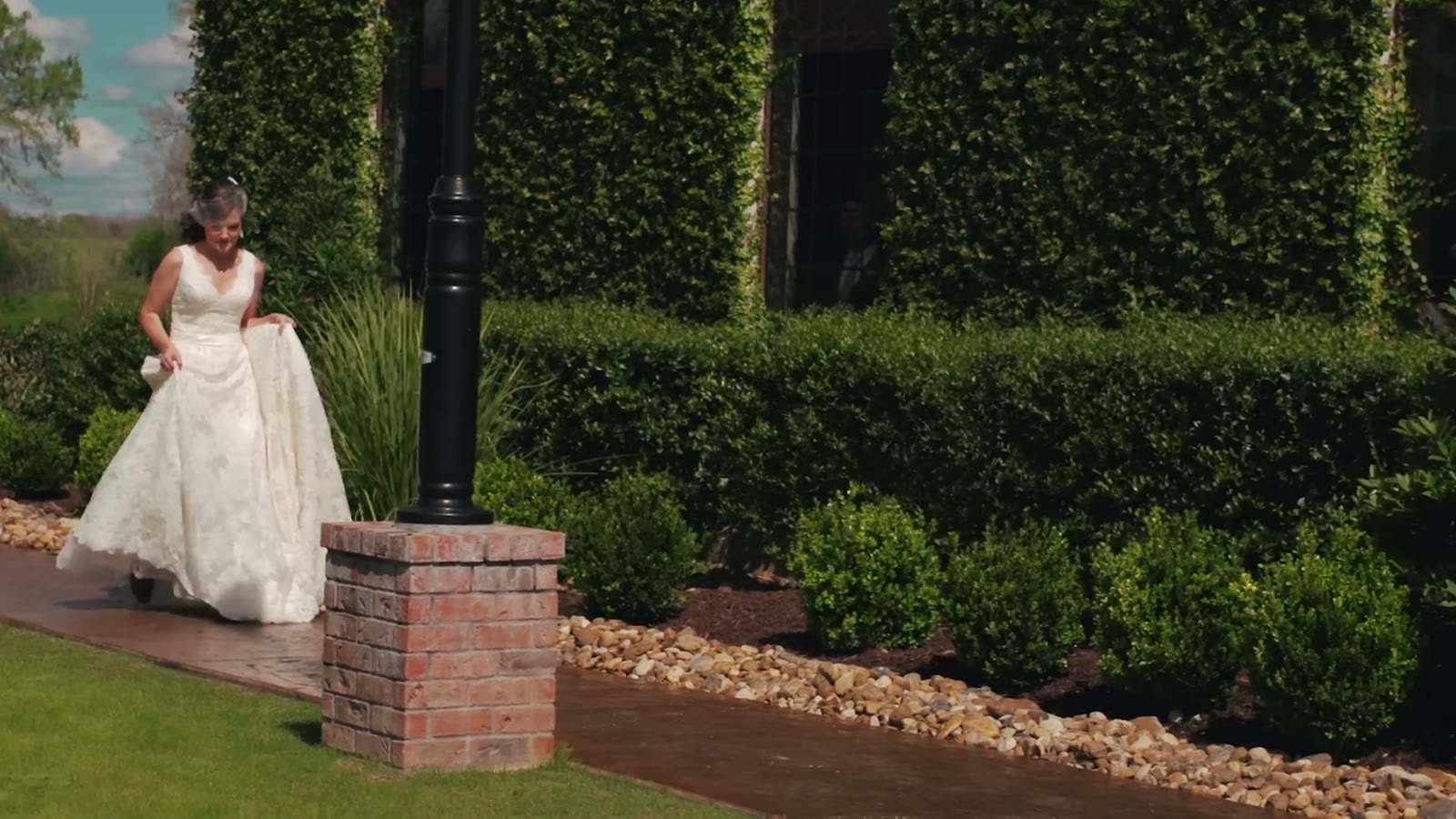Bride walking along a brick path lined with an ivy-covered wall, holding up the front of her dress