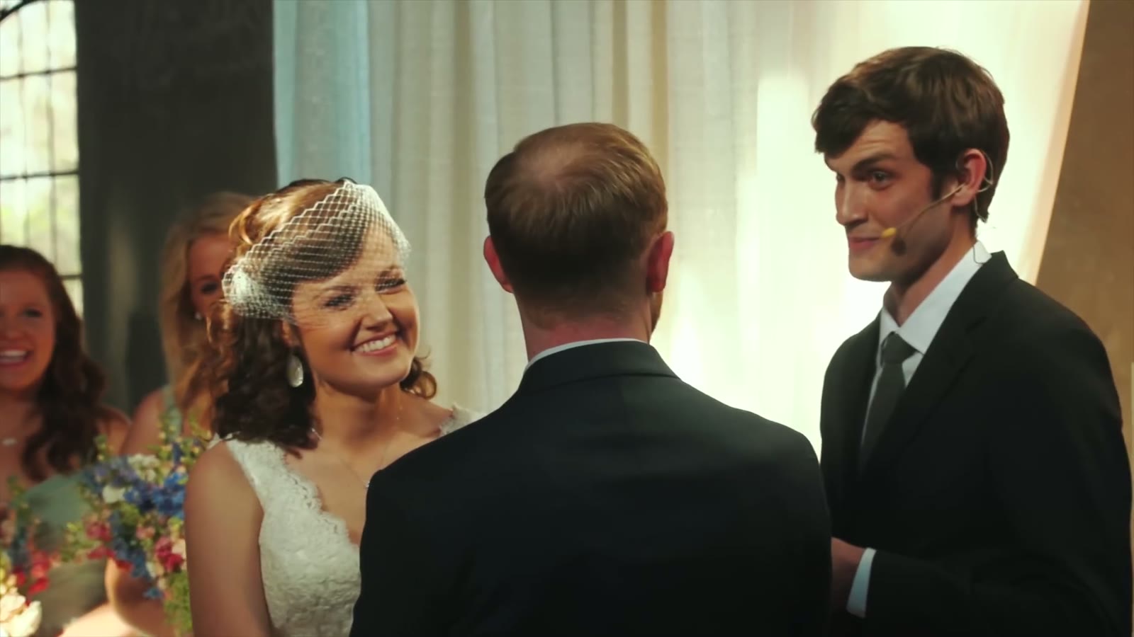 Bride in a birdcage veil laughing during the ceremony as the officiant with a headset mic looks on