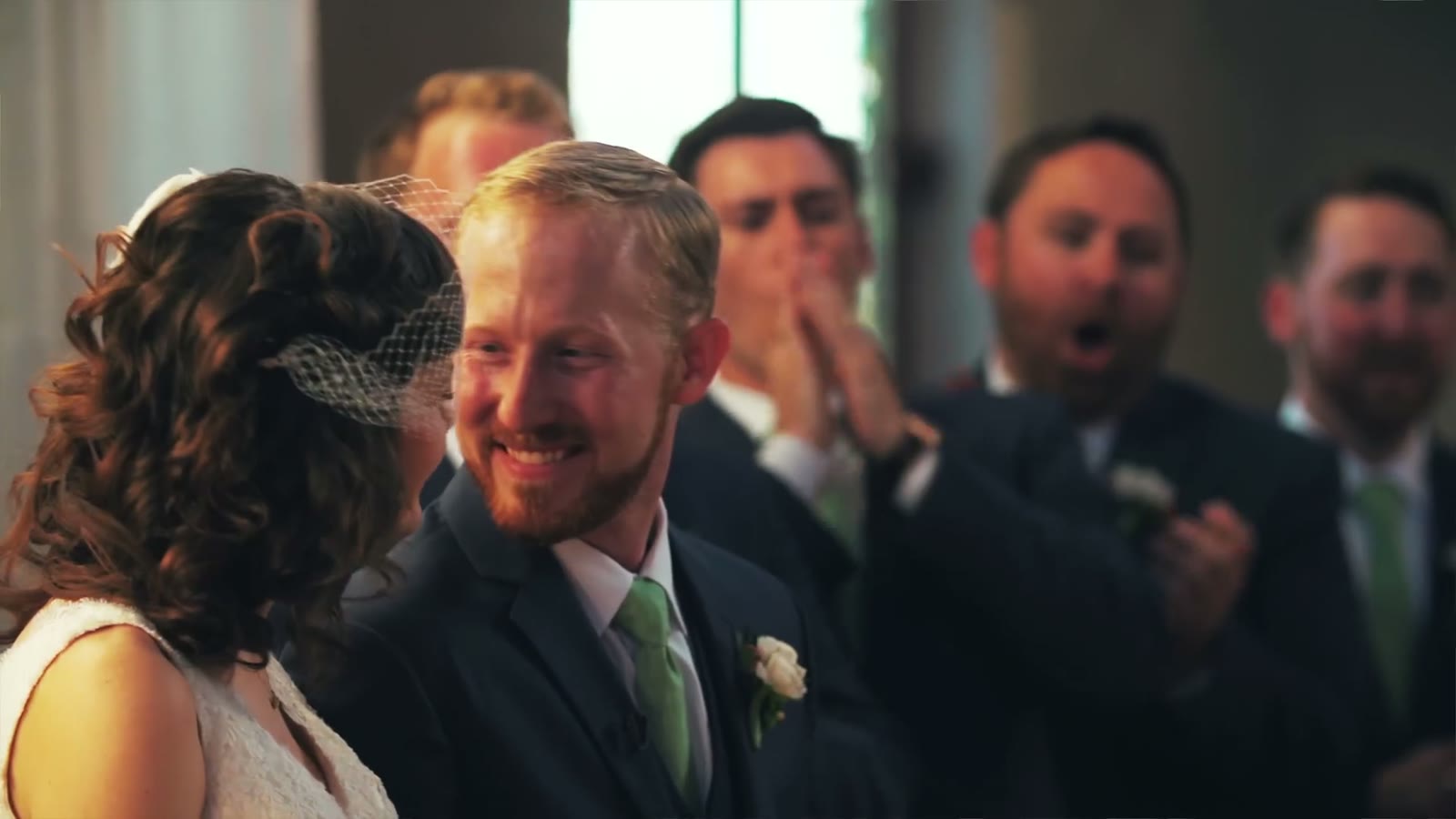Close shot on the groom smiling at the bride while groomsmen behind him laugh and clap