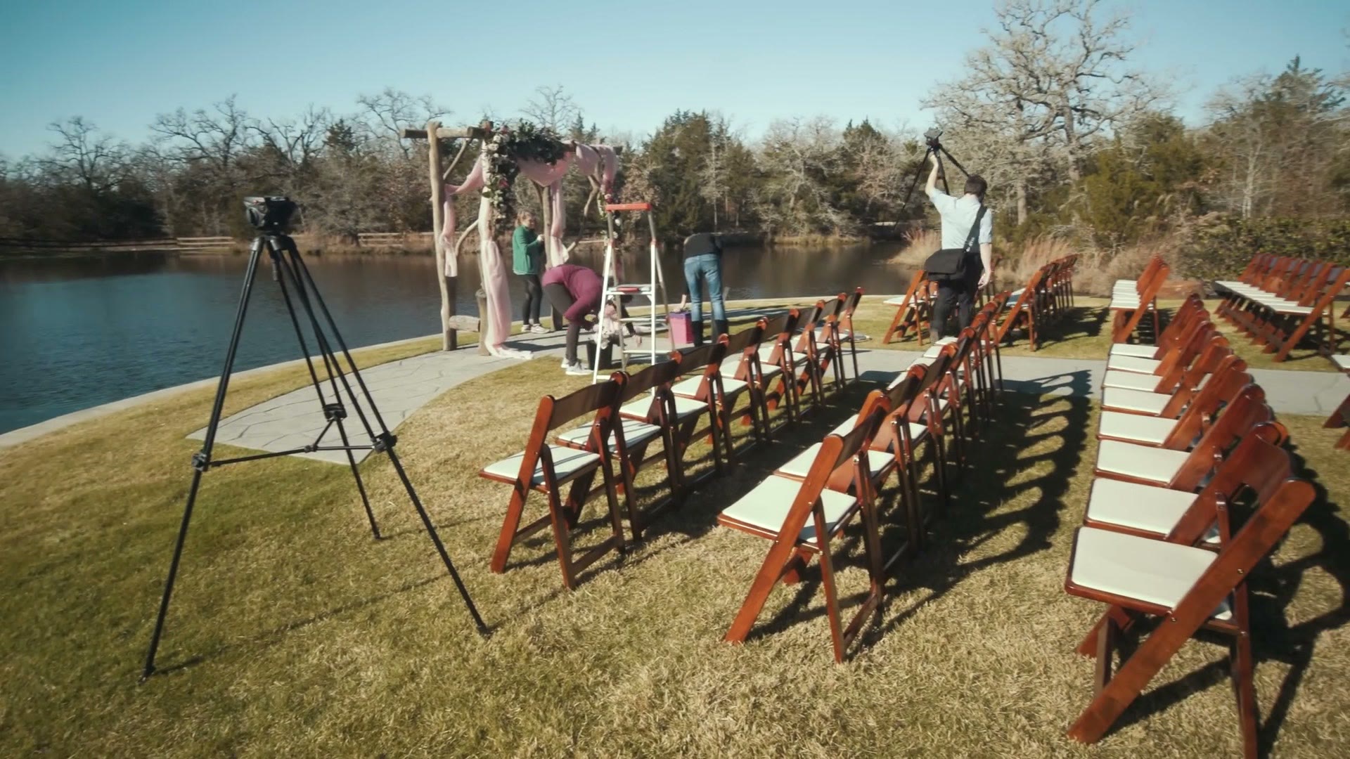 A tripod set up on the grass beside a lake, pointing at an outdoor ceremony arch while the venue team finishes decorating it.