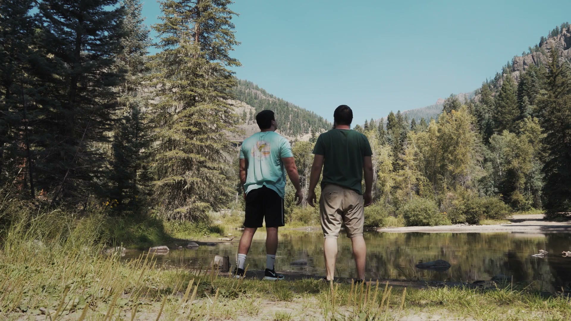 Two grown brothers standing side by side at the edge of a still pond, seen from behind, framed by pine trees and mountains