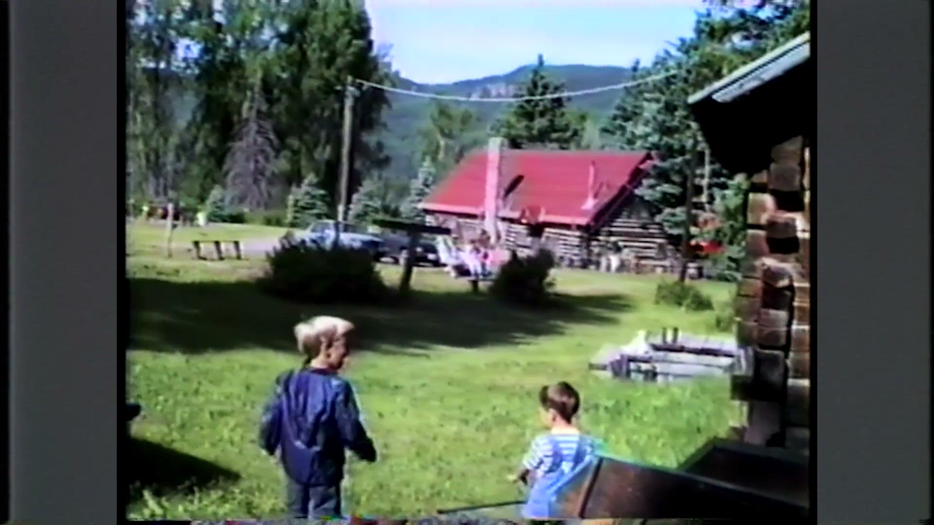 Camcorder footage from 1992 showing log cabins on a lawn with two small children playing in front of them