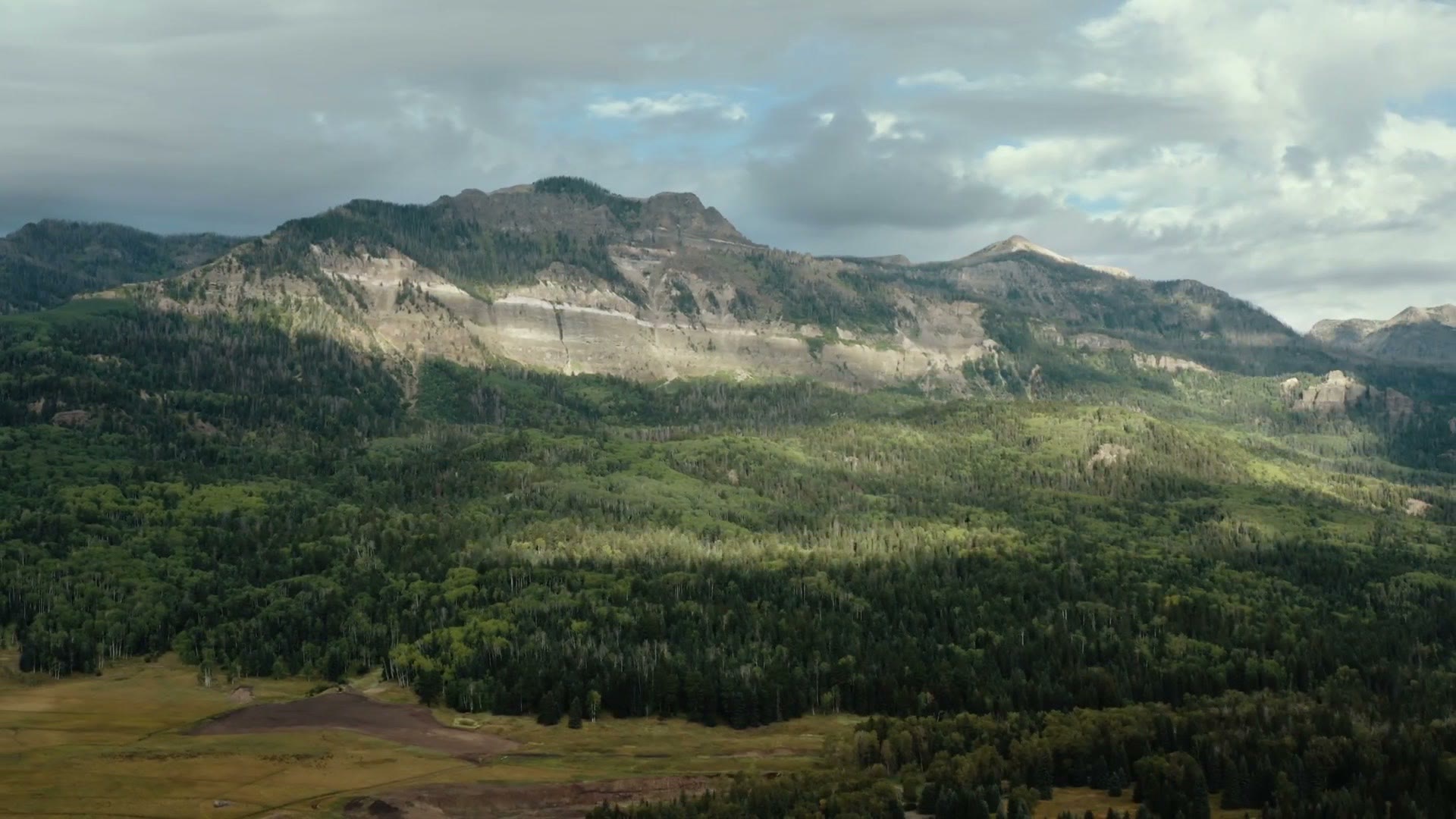 An aerial view over a forested Colorado valley with a rocky ridge line and clouds above it