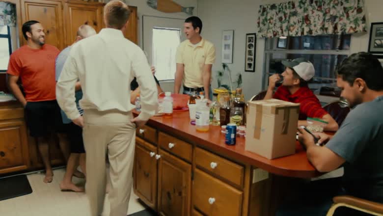 The groom getting dressed in a family kitchen with half a dozen relatives around him, drinks and a cardboard box on the counter