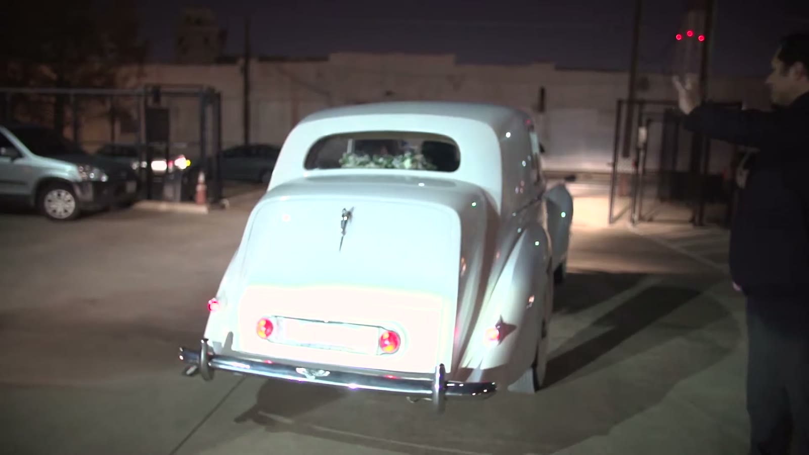 Vintage cream sedan pulling out of a parking lot at night, taillights glowing as a guest waves it off