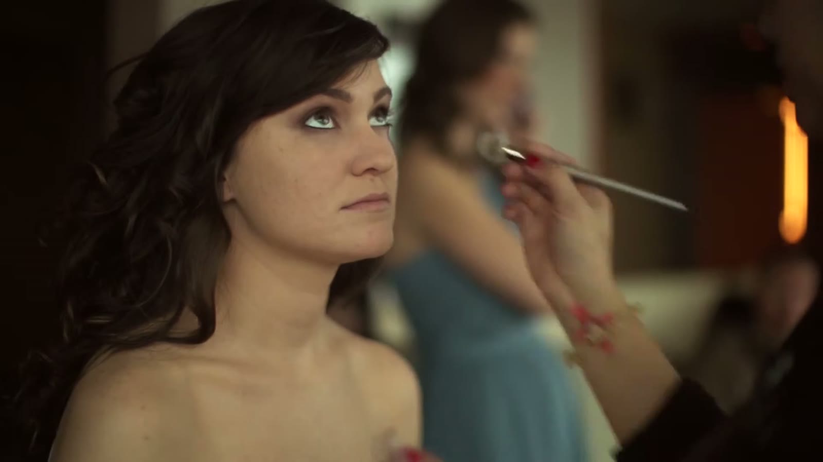 Bride having eye makeup applied, a bridesmaid in blue being styled softly out of focus behind her