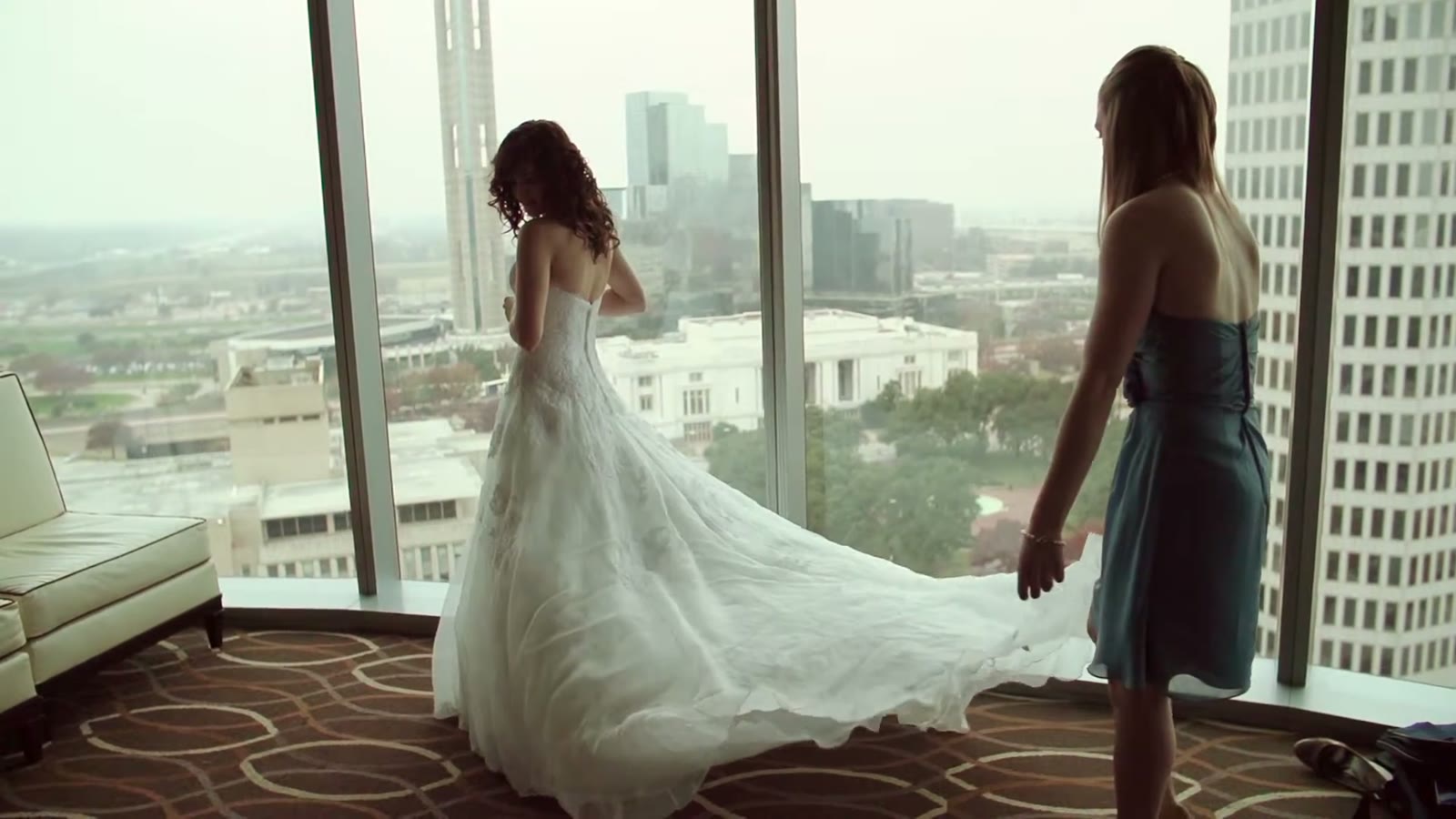 Bride in her gown looking out a floor-to-ceiling window at the Dallas skyline while a bridesmaid holds her train
