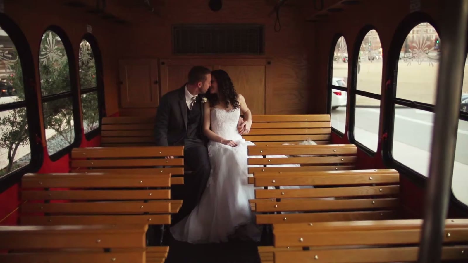 Bride and groom kissing on a wooden-bench trolley, city street visible through the arched windows