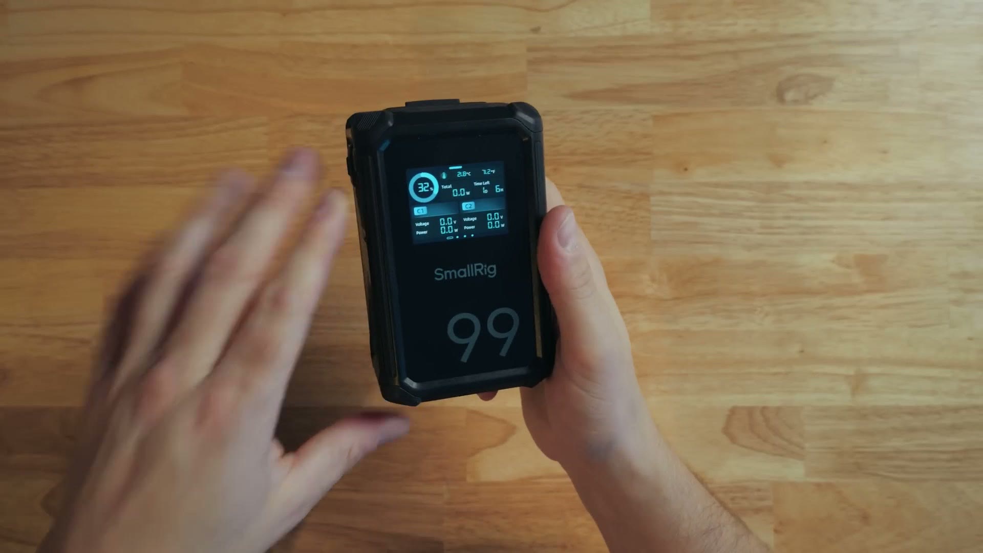 Hands holding the SmallRig VB99 Pro V-mount battery, its color touchscreen showing per-output power draw