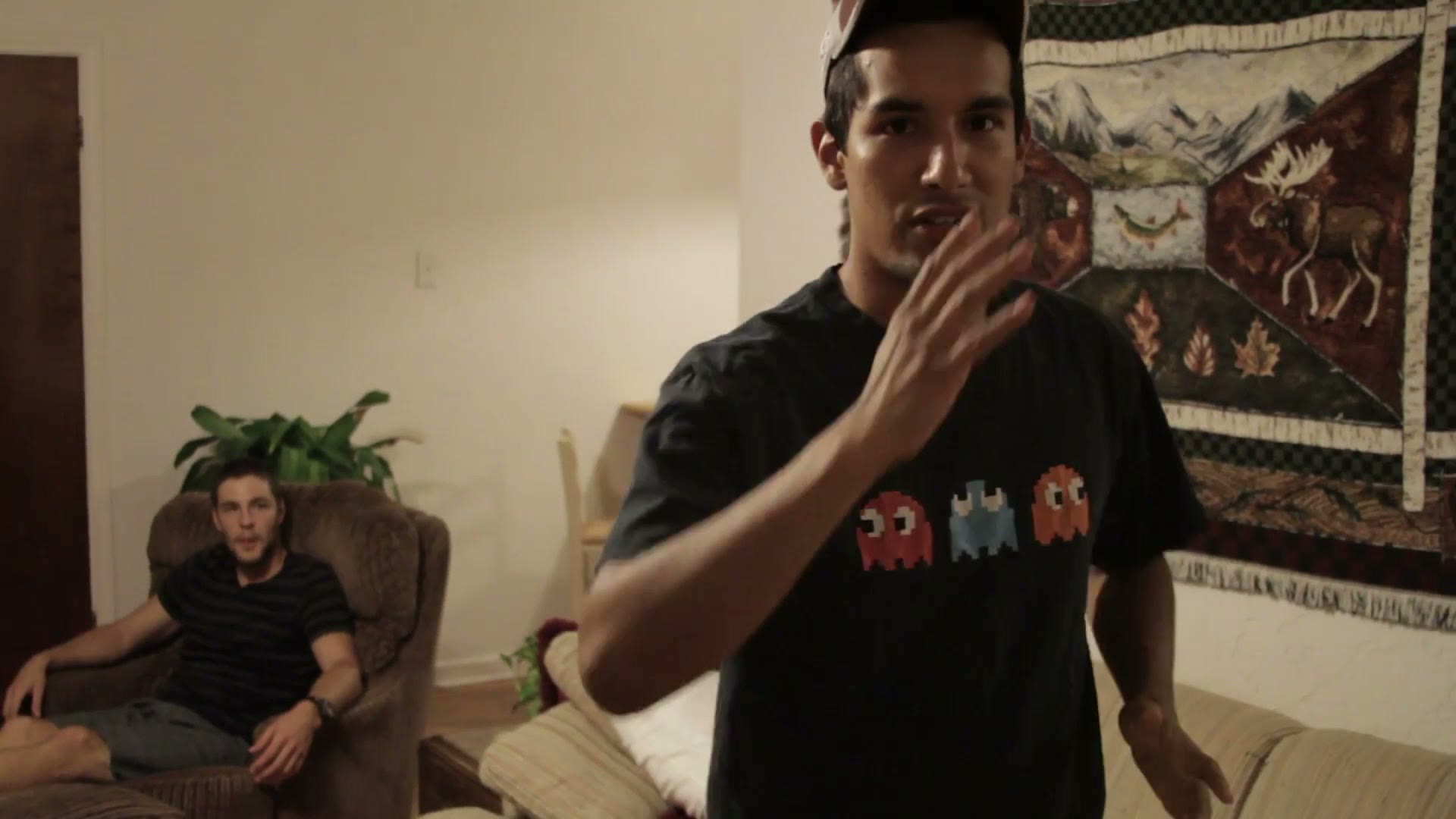 A young man standing in the living room gesturing with his hands as he describes a shot, with a patterned moose blanket hanging on the wall behind him
