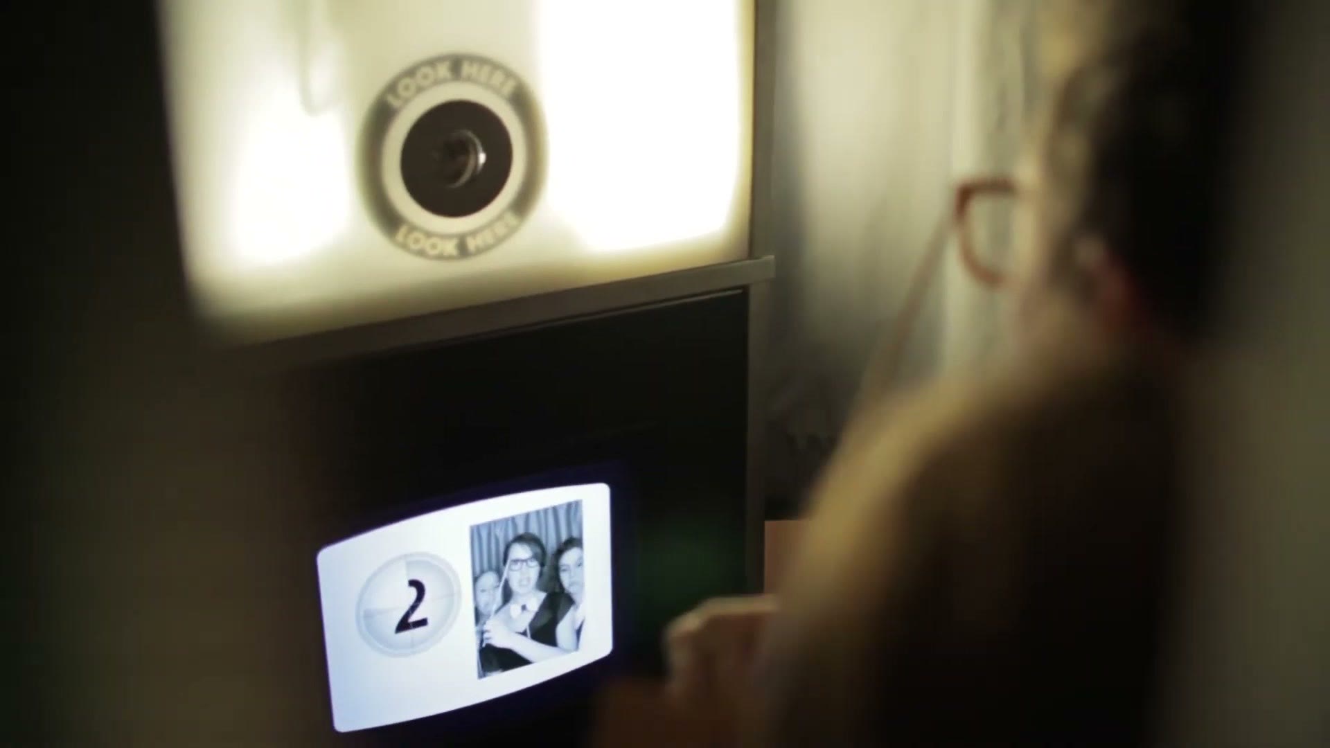 The inside of a photo booth showing the camera lens ringed with the words LOOK HERE, and a countdown on the screen below it