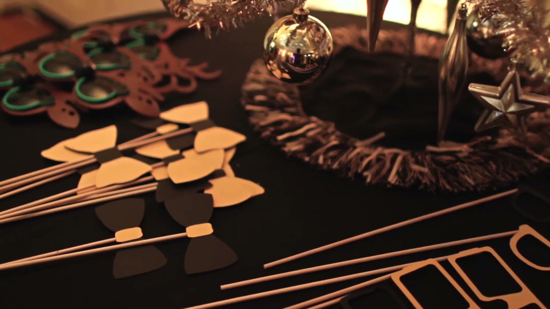 Gold paper bowties and glasses on sticks laid out on a black table beside Christmas baubles and a tinsel wreath