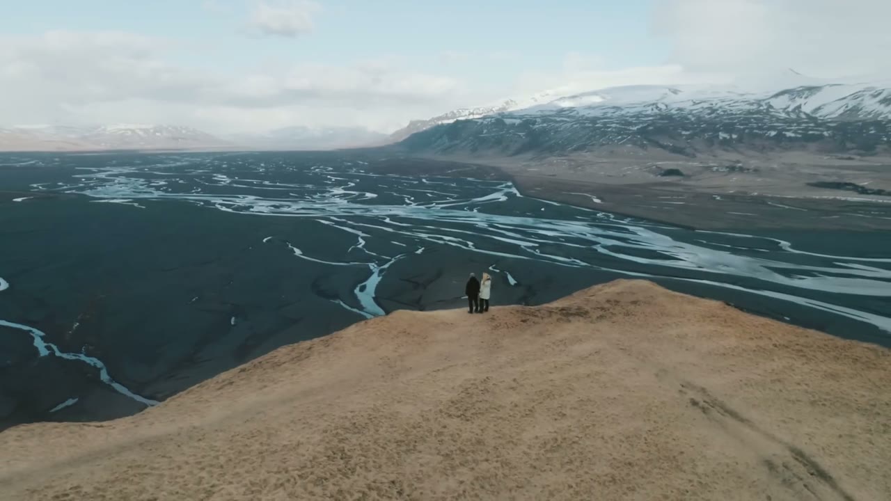 Aerial view of a couple standing on a ridge overlooking braided glacial rivers cutting through black sand