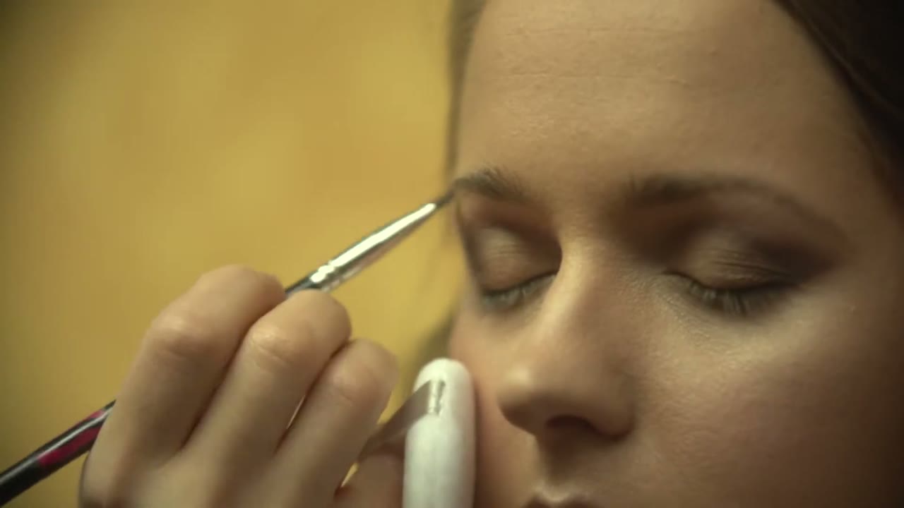 Close-up of a makeup artist applying eyeshadow to the bride's closed eye against a warm gold-lit background