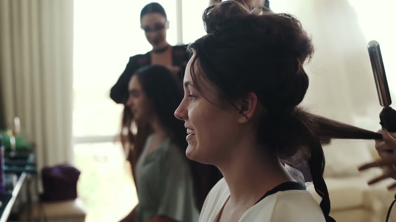 Bride smiling while a stylist curls her hair, other bridesmaids visible in soft window light behind her