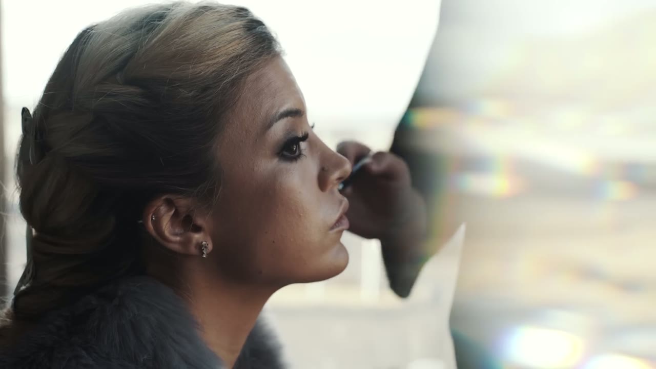 Woman getting her makeup done in profile, a mascara wand near her eye, soft window light behind her