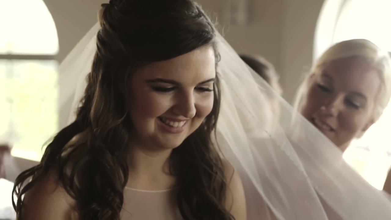 Bride laughing while a bridesmaid arranges her veil from behind, arched windows in the background