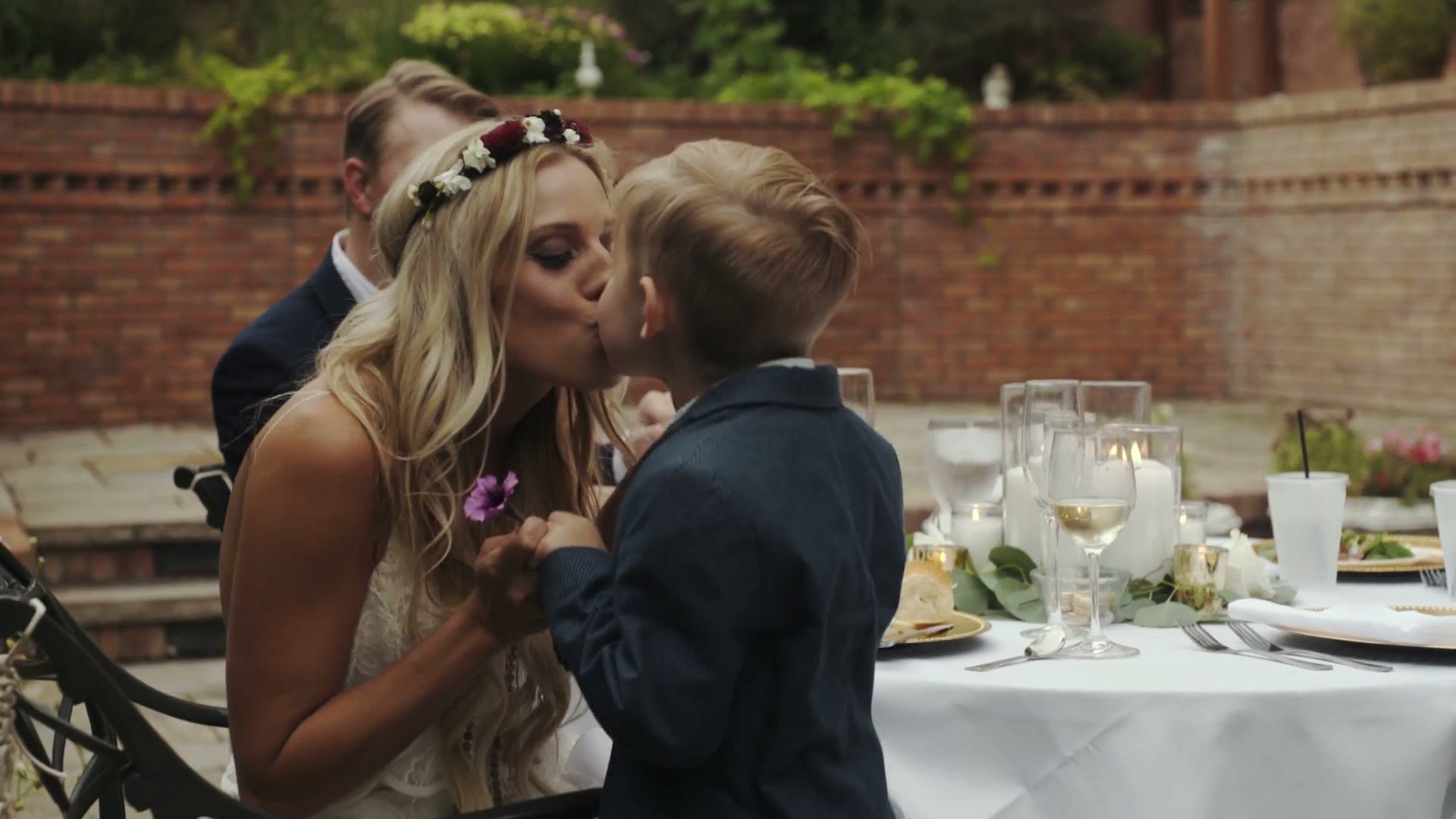 Bride kissing her young son on the cheek at the outdoor reception table, groom blurred behind them