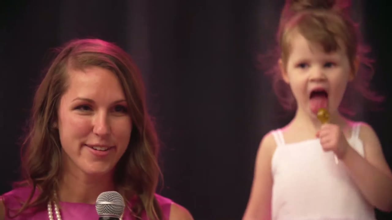 Woman speaking into a microphone during toasts while a flower girl in white licks a lollipop beside her