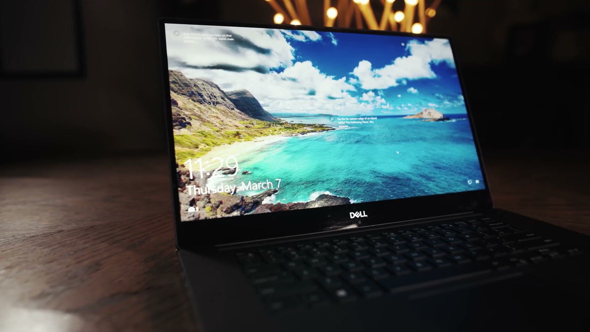 A Dell XPS 15 open on a wooden desk in a dark room, its lock screen lit up, with warm fairy lights out of focus behind it.