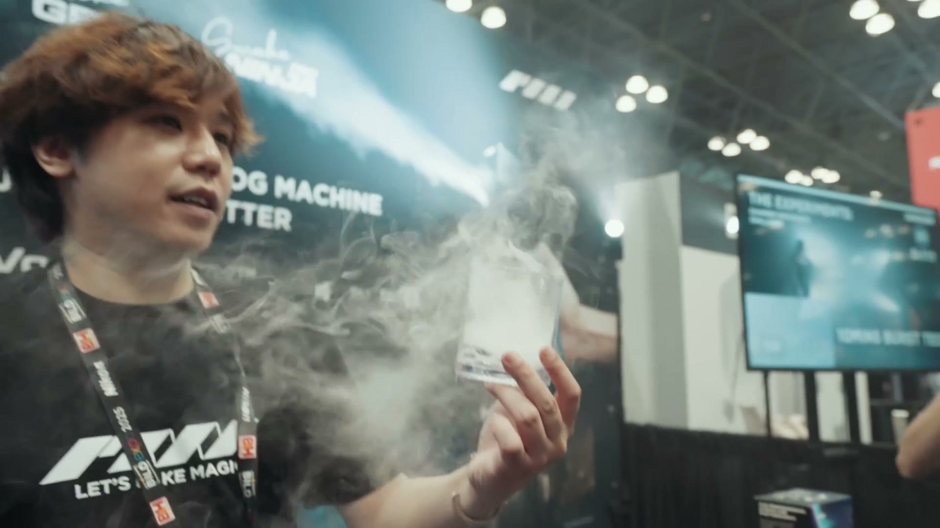 A handheld fog machine being demonstrated at a booth, throwing a cloud of white fog across the frame