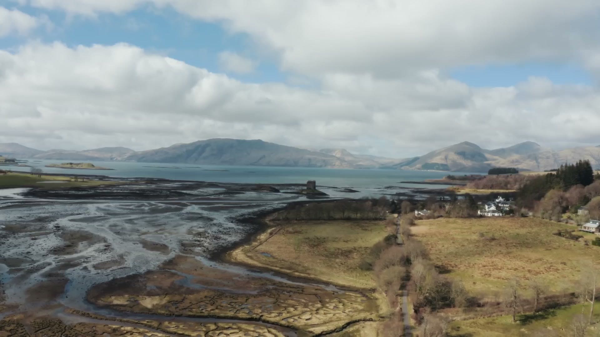 Aerial drone view of Castle Stalker sitting on a small island in a tidal loch, with mountains across the water and blue sky above
