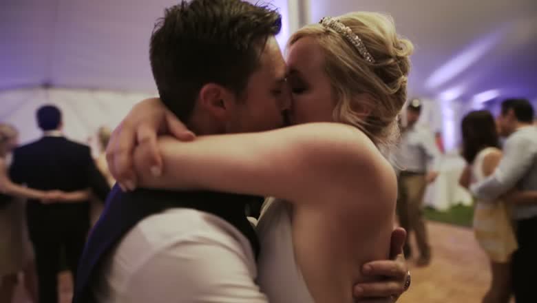 The couple kissing on the dance floor inside the white tent, guests dancing around them under purple uplighting