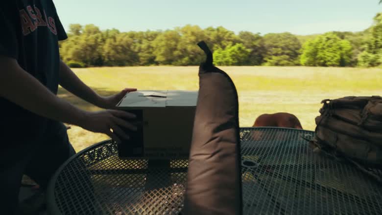 Two men setting up a clay thrower and a box of shells on a garden table beside a gun case, with an open field behind