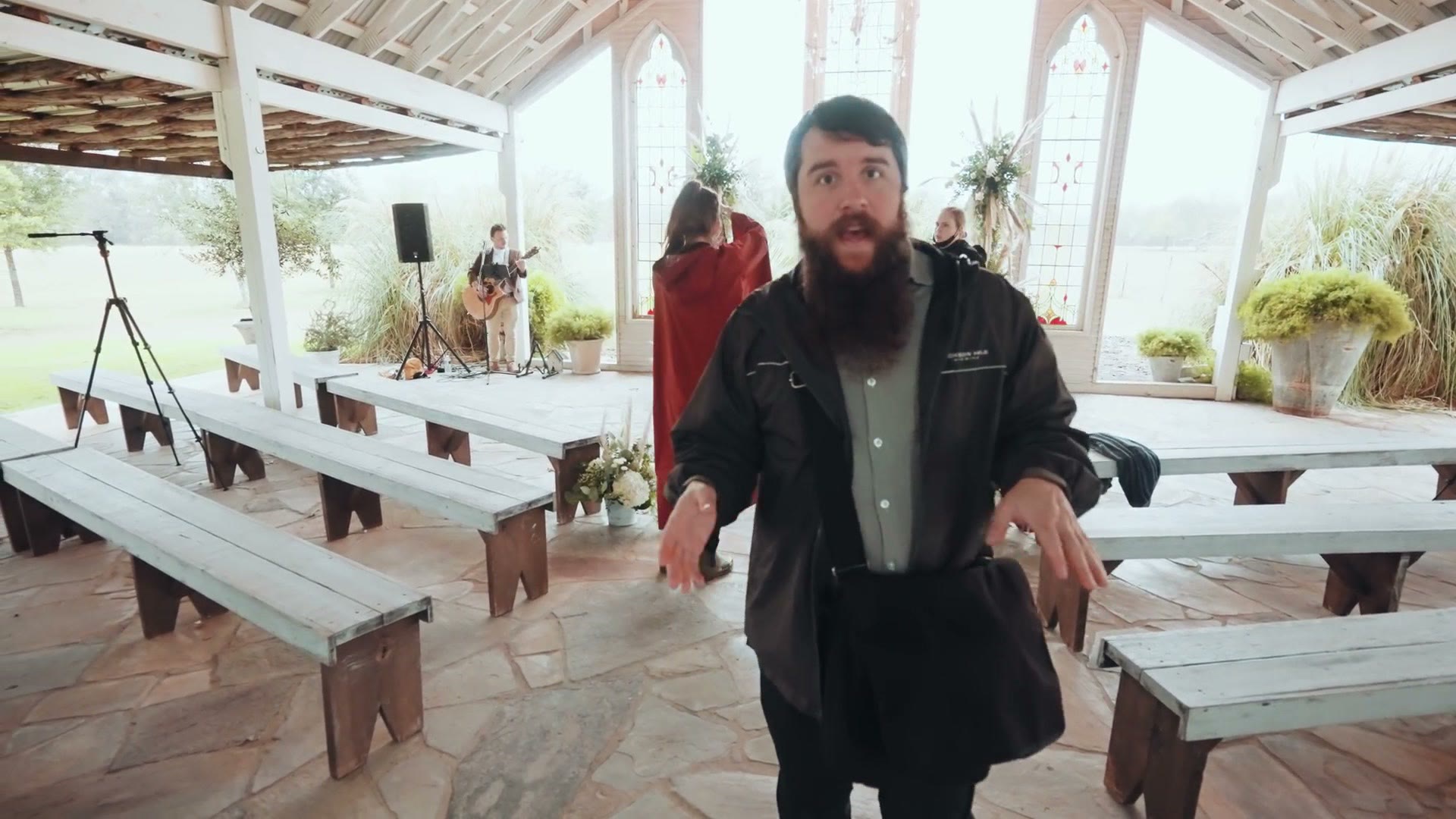 Matt, bearded and gesturing, talking to camera in an empty open-air wooden chapel with a guitarist behind him