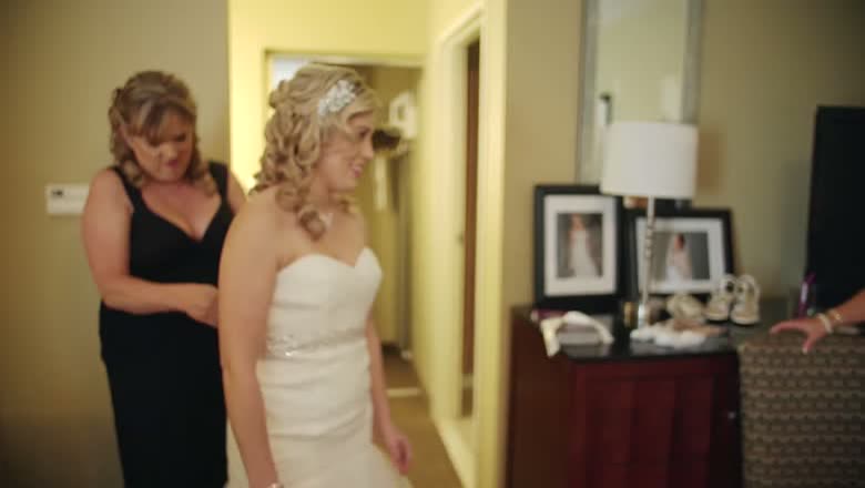 The bride standing in her dress while her mother fastens the back of it in a getting ready room