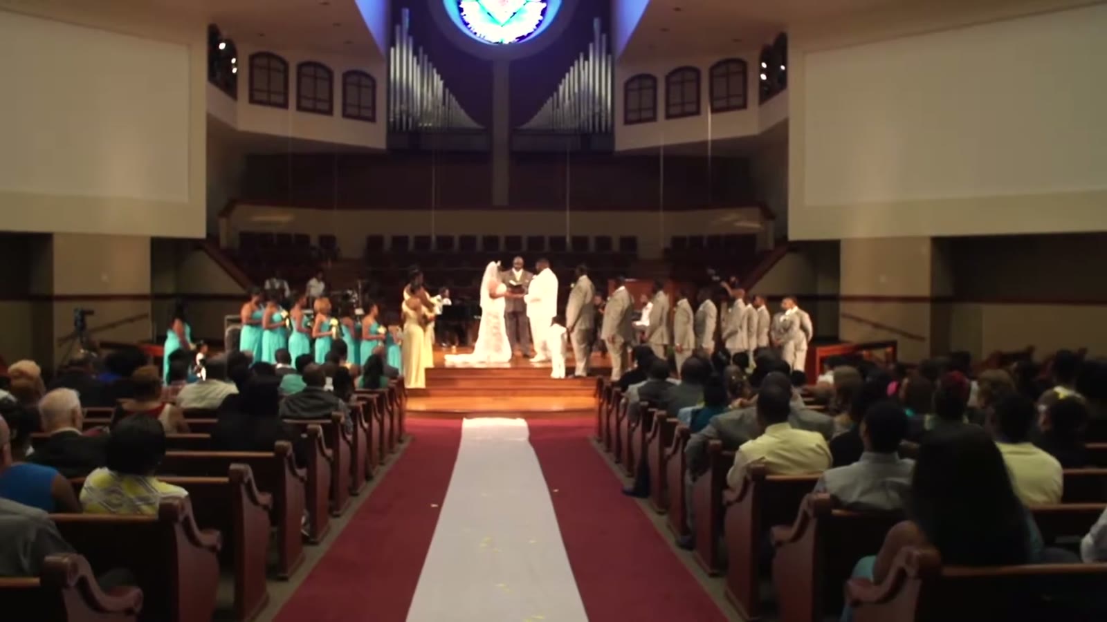 Wide view of the church sanctuary during the ceremony, wedding party on stage and guests filling the pews along a red aisle runner