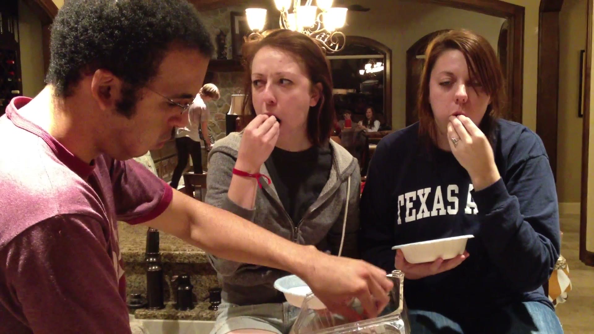 Two young women sitting at a kitchen counter each holding a white bowl, both putting grapes into their mouths, with a friend refilling a container in front of them