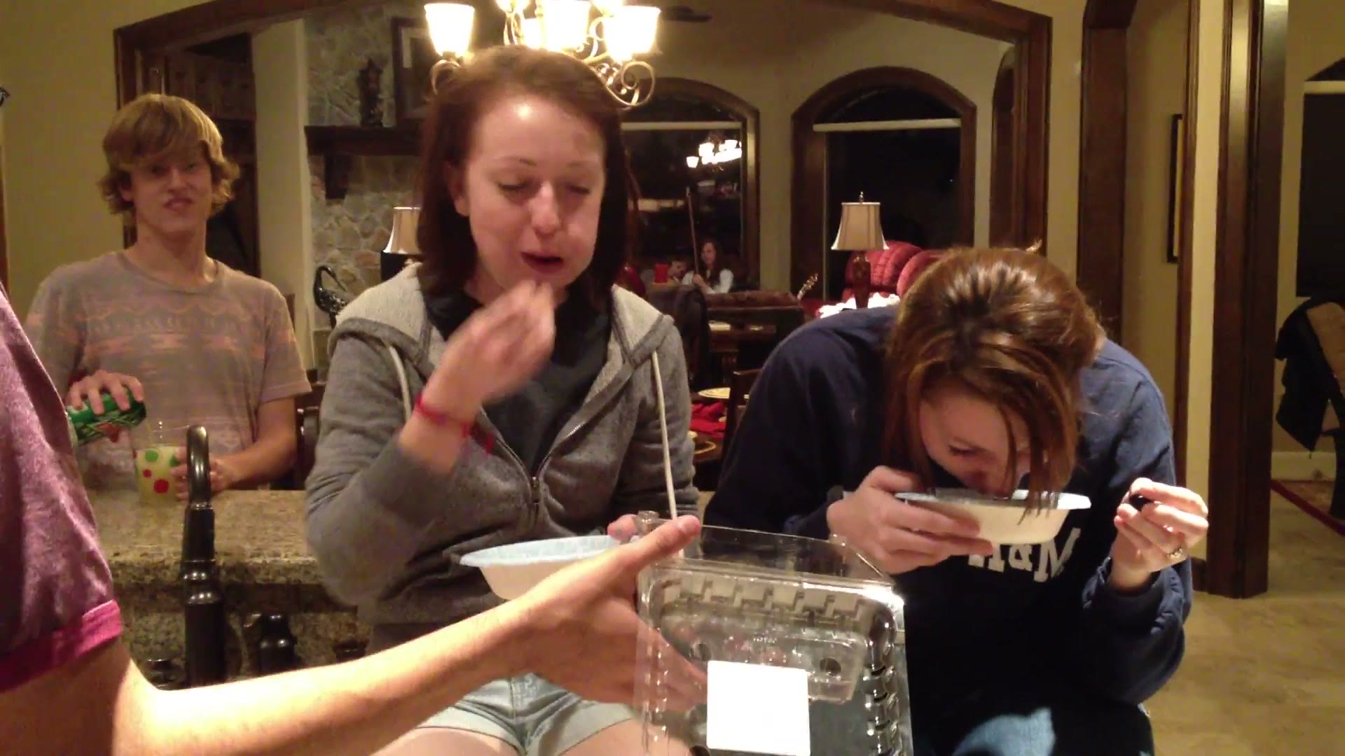One competitor grimacing with full cheeks while the other leans forward over her bowl, with party guests visible in the background