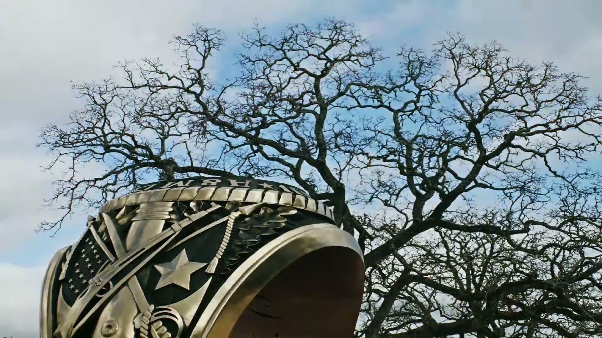 A large bronze sculpture of an Aggie Ring photographed from below against the bare branches of a live oak and a blue sky