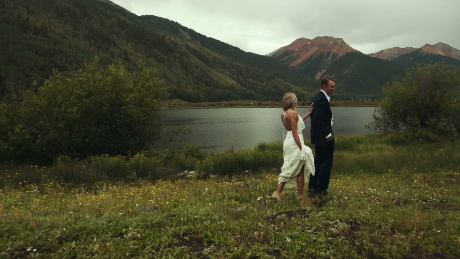 Bride and groom walking beside a mountain lake with jagged Colorado peaks and wildflowers in the foreground