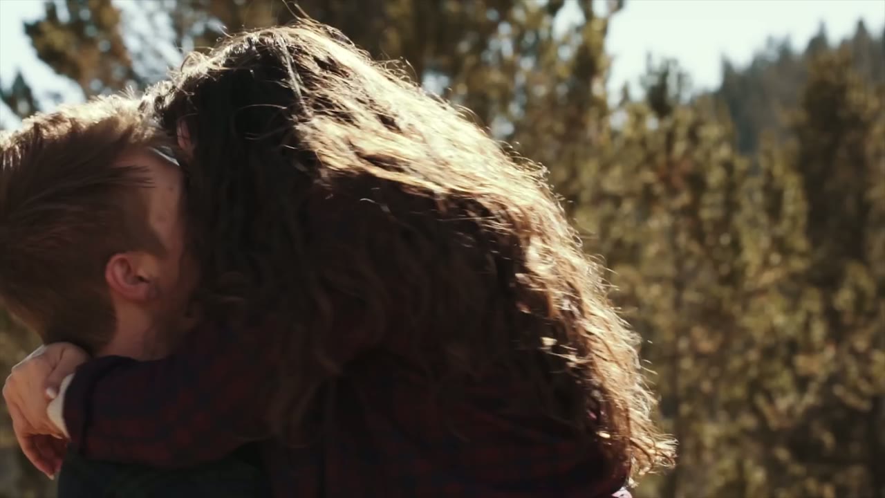 Close-up of the couple kissing outdoors, her hair backlit against a pine forest