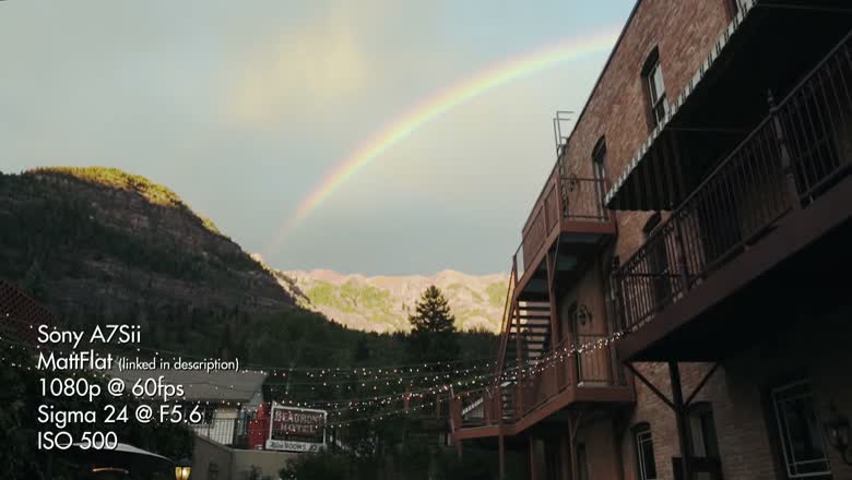 A rainbow over the mountain behind a hotel balcony strung with lights, with a burned-in settings overlay reading Sony A7Sii, MattFlat, 1080p at 60fps, Sigma 24 at F5.6, ISO 500