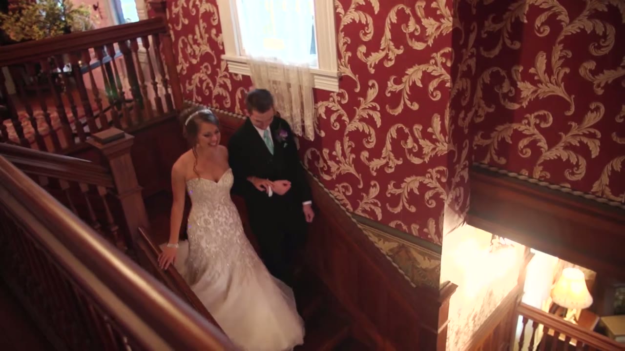 Bride and groom descending an ornate wooden staircase past red patterned wallpaper inside the Calvert Inn