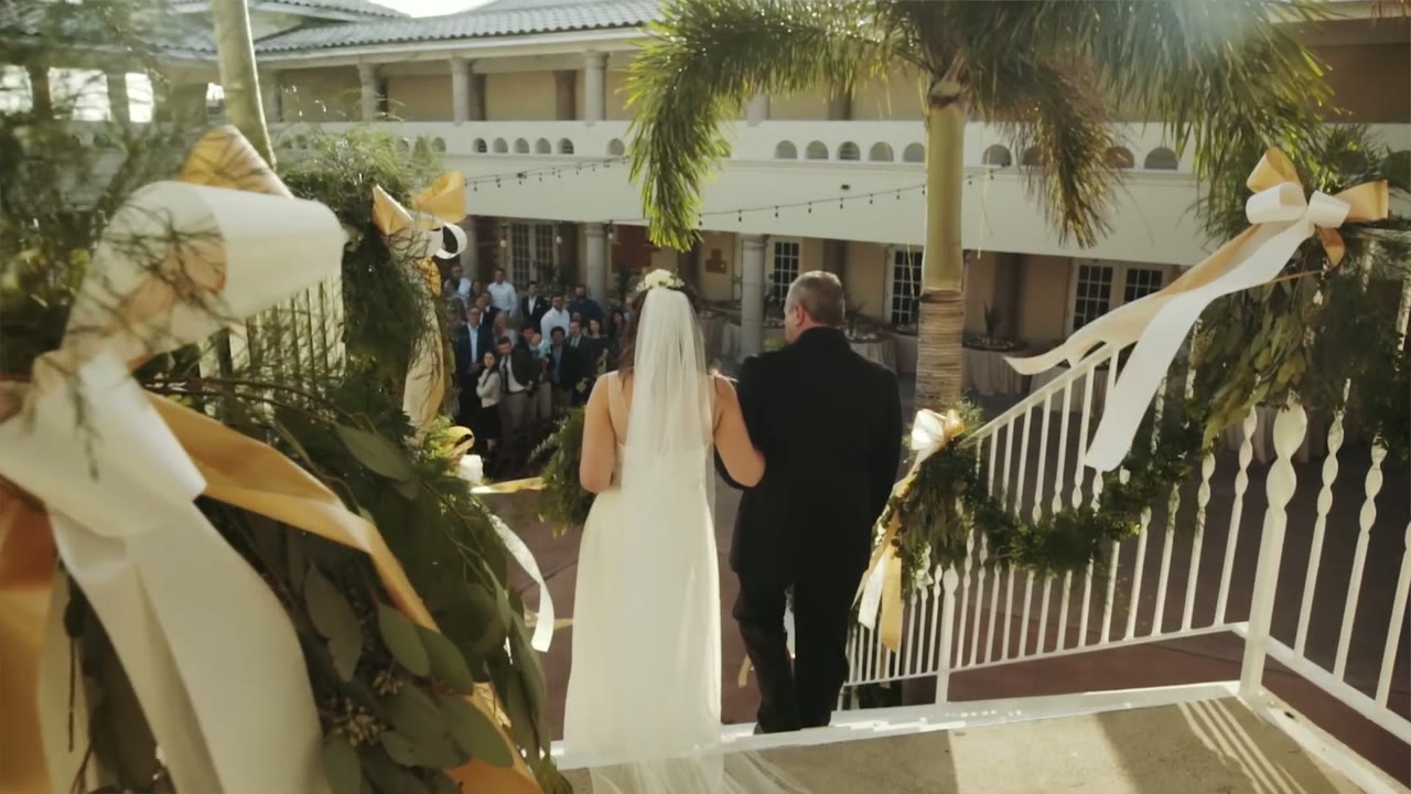 Bride and groom walking down palm-lined steps decorated with gold ribbon garlands toward a courtyard of guests