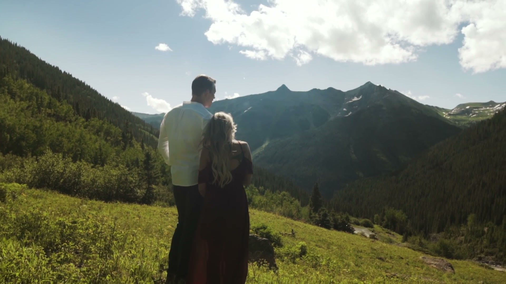 Couple seen from behind standing on a hillside looking out over a green valley and mountain range